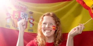A smiling young woman holding a small Spanish flag in a vibrant Madrid street, illustrating the lifestyle and cultural integration for English teachers in 2026.