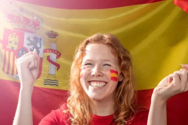A smiling young woman holding a small Spanish flag in a vibrant Madrid street, illustrating the lifestyle and cultural integration for English teachers in 2026.
