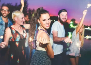 A diverse group of smiling friends celebrating outdoors at sunset with sparklers, capturing a vibrant and social atmosphere.