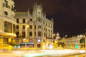 Long exposure of the Plaza de Cibeles area at night, showing the illuminated classical architecture and light trails from the traffic.