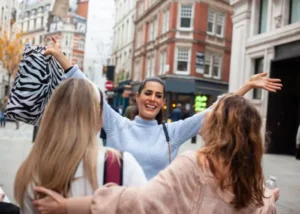 Three young women smiling and greeting each other with open arms on a Madrid city street, representing a warm reunion and friendship.