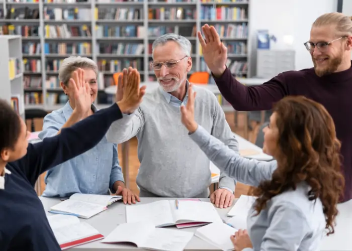A diverse group of adult students and their teacher giving a high five over a desk with open books in a library, celebrating a shared achievement.