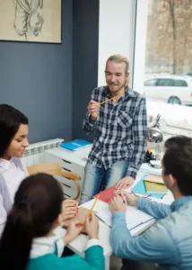 A young male teacher in a plaid shirt smiling and holding a pencil while leading a discussion with a group of diverse students at a table in a bright, modern classroom.