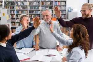 A diverse group of adult students and their teacher giving a high five over a desk with open books in a library, celebrating a shared achievement.