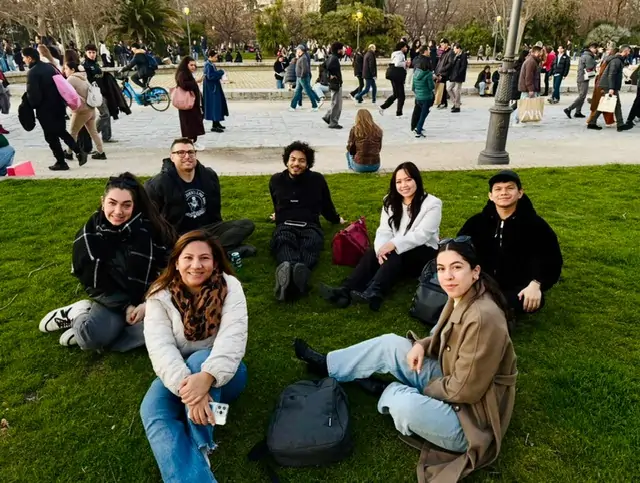 A group of young adults, including Joanna, sitting on the green grass of a Madrid park with a fountain and many people in the background.