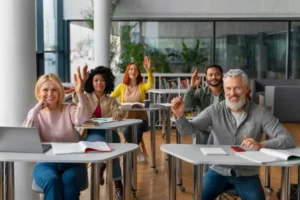 A diverse group of adult students sitting at individual desks in a bright, modern classroom, all smiling and raising their hands with enthusiasm.