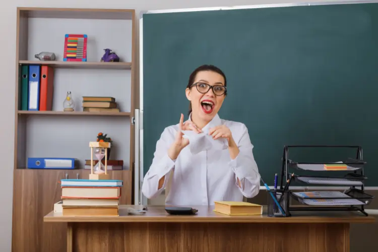 A young female teacher wearing glasses and a white shirt, sitting at her desk in front of a blackboard, tearing a piece of paper with an expression of crazy happiness and triumph.