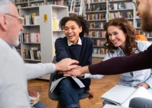 A diverse group of adult students sitting at individual desks in a bright, modern classroom, all smiling and raising their hands with enthusiasm during a lesson.
