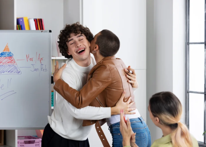 A student in a white sweater being hugged by a peer in a brown jacket next to a whiteboard, while another student claps in the foreground.