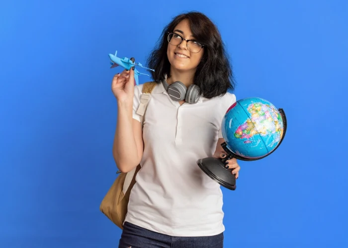 A young woman holding a small globe and a toy airplane, looking upward with a hopeful expression as if dreaming of international travel and a new life abroad.