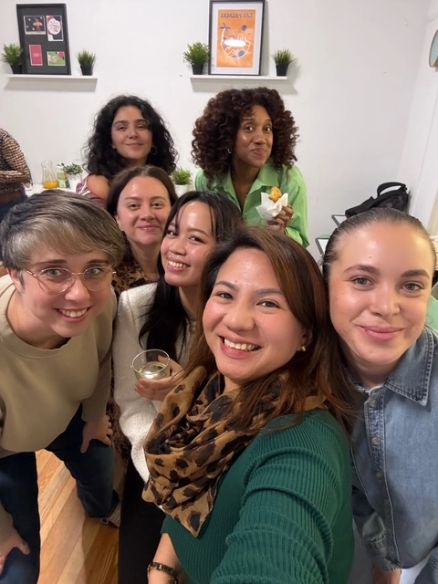 A high-angle group selfie of smiling students, including Joanna in a green top, enjoying a social gathering indoors.