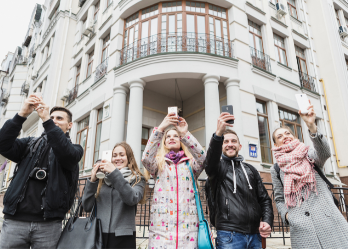 A diverse group of young adults standing in front of a white Madrid building, smiling and holding up their phones.