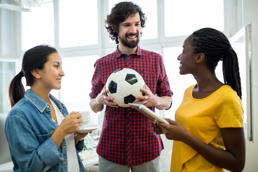 A diverse group of three young professionals smiling and talking during a coffee break, with a man in a red plaid shirt holding a football in the center.