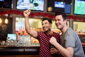 Two cheerful men celebrating and raising their fists while holding beer bottles at a sports bar with television screens showing a football match in the background.