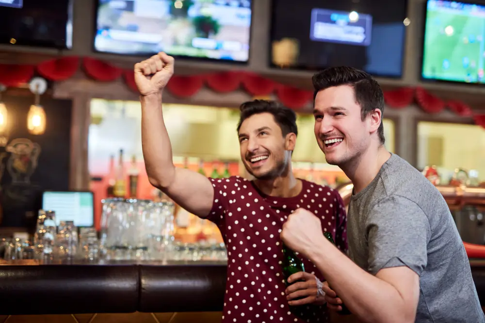 Two cheerful men celebrating and raising their fists while holding beer bottles at a sports bar with television screens showing a football match in the background.