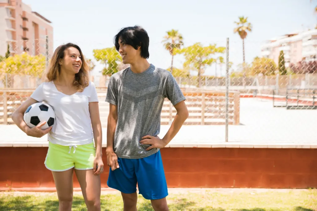 A young woman holding a soccer ball under her arm and a young man smiling and laughing together on an outdoor sports field on a sunny day.