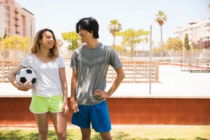 A young woman holding a soccer ball under her arm and a young man smiling and laughing together on an outdoor sports field on a sunny day.