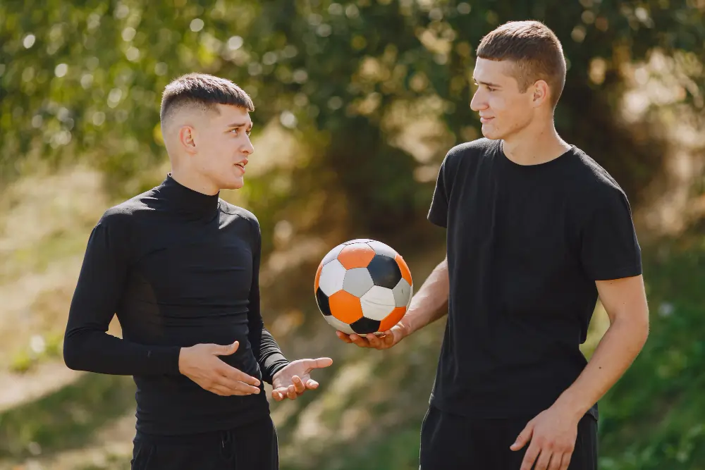 Two young men in black athletic shirts standing outdoors, deeply engaged in a passionate conversation while one holds an orange and black football.