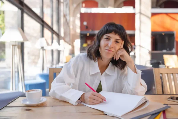 A young woman sitting at a sunny wooden table in a spacious Madrid café, smiling thoughtfully while writing in a notebook next to a cup of coffee.