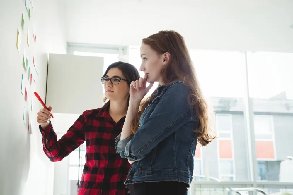 A professional career coach with glasses pointing to notes on a wall during a one-on-one session with a student inside a bright TtMadrid classroom.