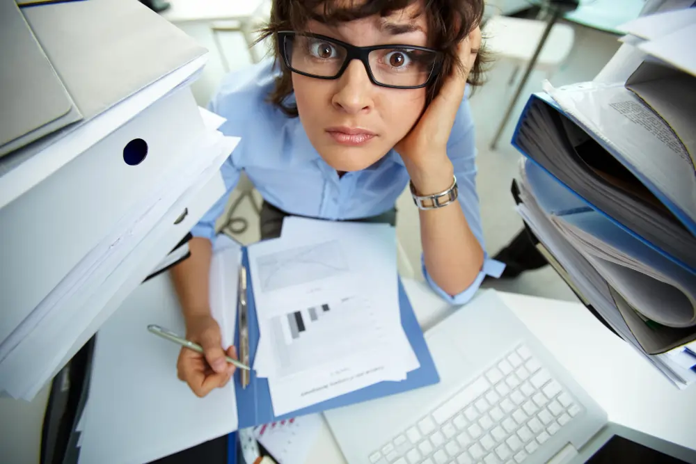 A wide-angle top-down view of a wide-eyed, stressed woman wearing glasses at a desk surrounded by massive, towering stacks of white paperwork and folders.