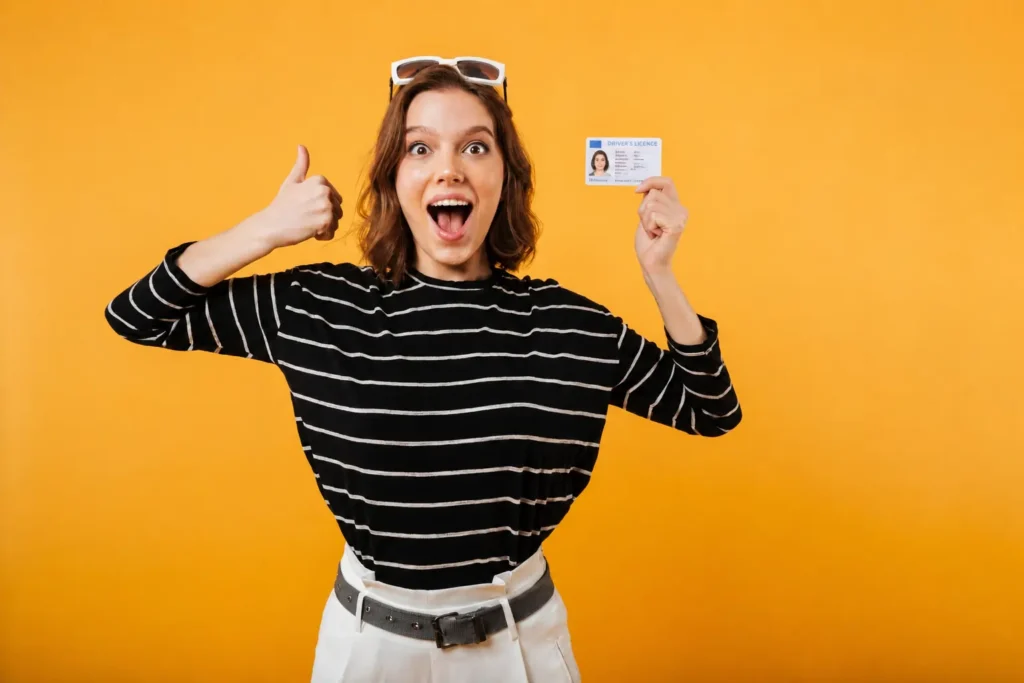 An excited, smiling young woman holding up an identity card in one hand and giving a thumbs-up with the other against a bright yellow background.