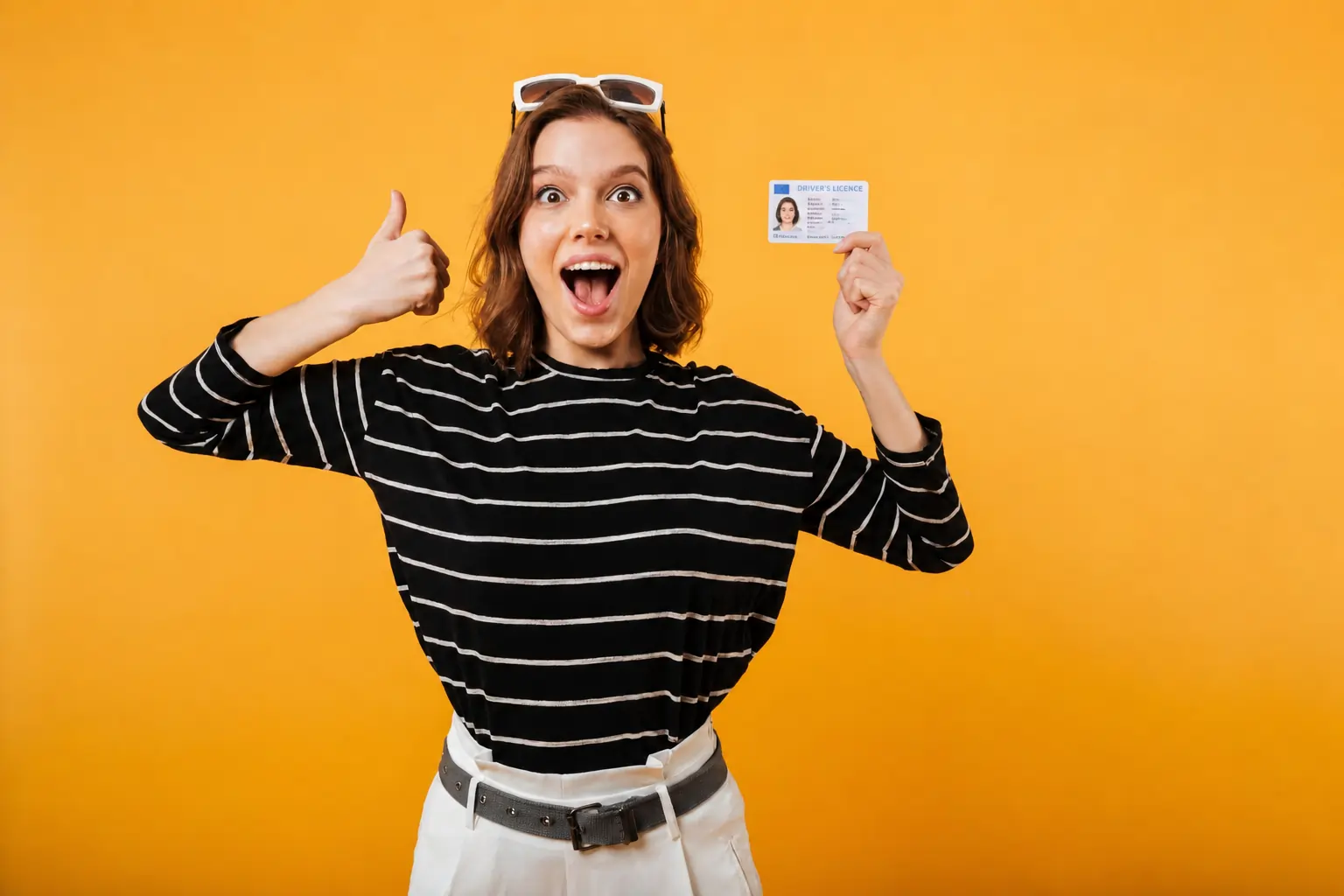 An excited, smiling young woman holding up an identity card in one hand and giving a thumbs-up with the other against a bright yellow background.